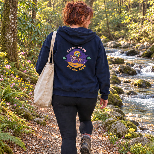 A woman viewed from behind walking on a forest path beside a flowing stream, wearing a navy hoodie with a large circular graphic on the back that reads 'Feral Moms Social Club' and carrying a beige tote bag.