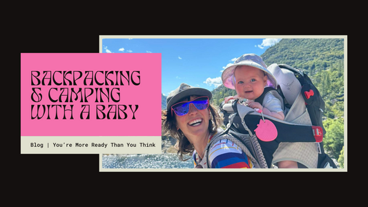 Smiling woman wearing sunglasses and a hat carries a baby in a hiking backpack on a sunny trail near a lake. The baby, also wearing a sunhat, looks at the camera. Text reads: 'Backpacking & Camping with a Baby – Blog | You’re More Ready Than You Think.'