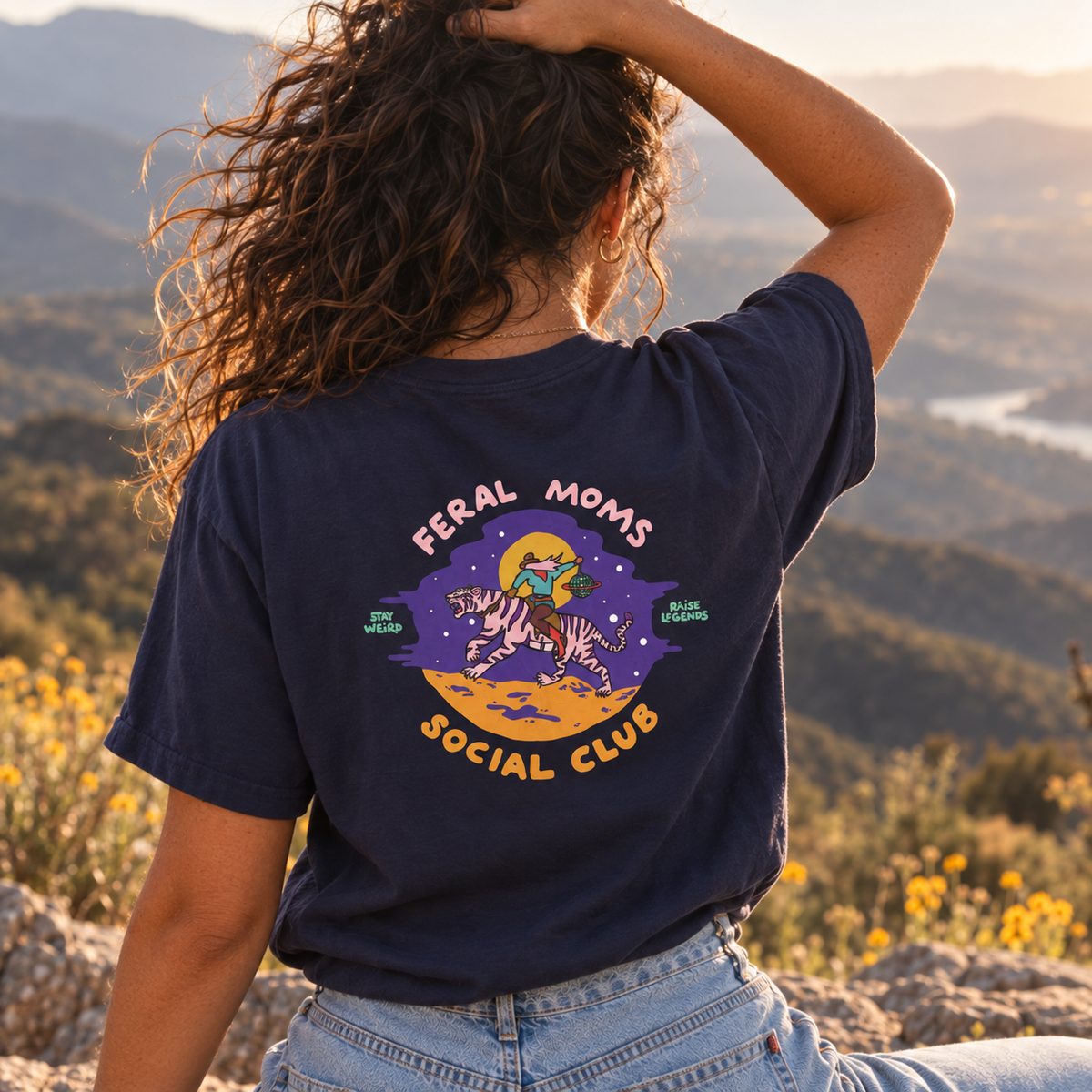 A woman seen from behind wearing a navy blue graphic t-shirt with a colorful 'Feral Moms Social Club' design on the back, set against a scenic outdoor background of mountains and wildflowers.