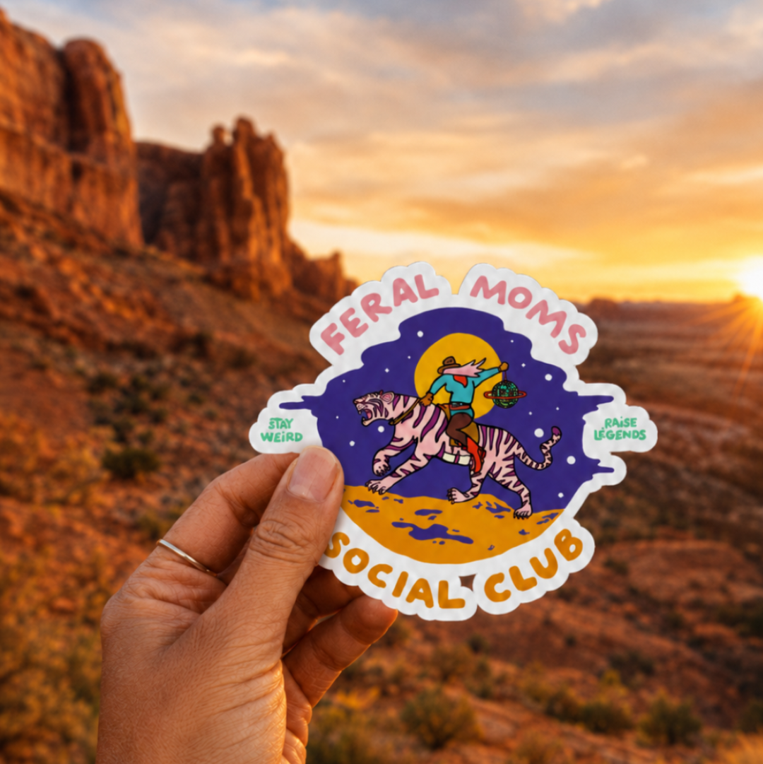Sticker with 'Feral Moms Social Club' design held against a desert sunset.