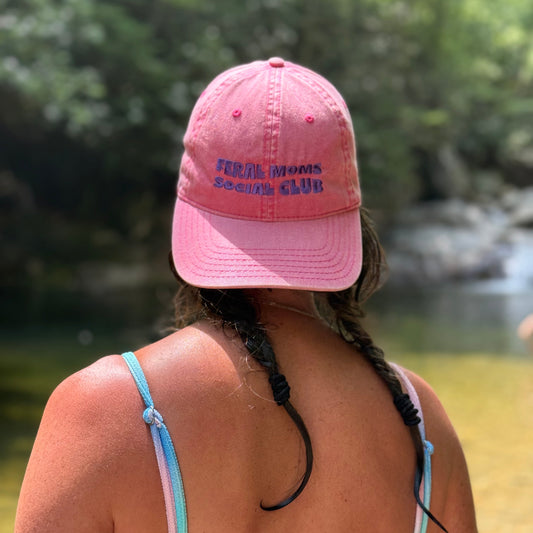 A woman viewed from behind wearing a pink baseball cap with purple embroidered text reading 'FERAL ANIMALS Social Club' and a pastel bikini at a sunny outdoor location by the water.