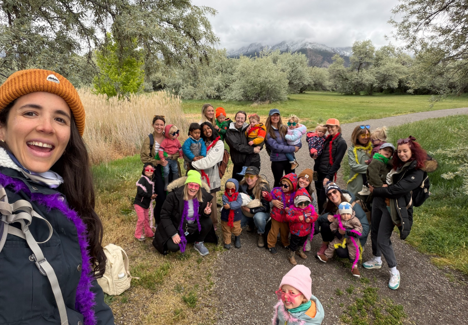 A large group of people posing together outdoors on a gravel path in a grassy park with trees and mountains in the background. The photo is taken in a casual group selfie style, with one woman in the foreground wearing an orange beanie and dark jacket.