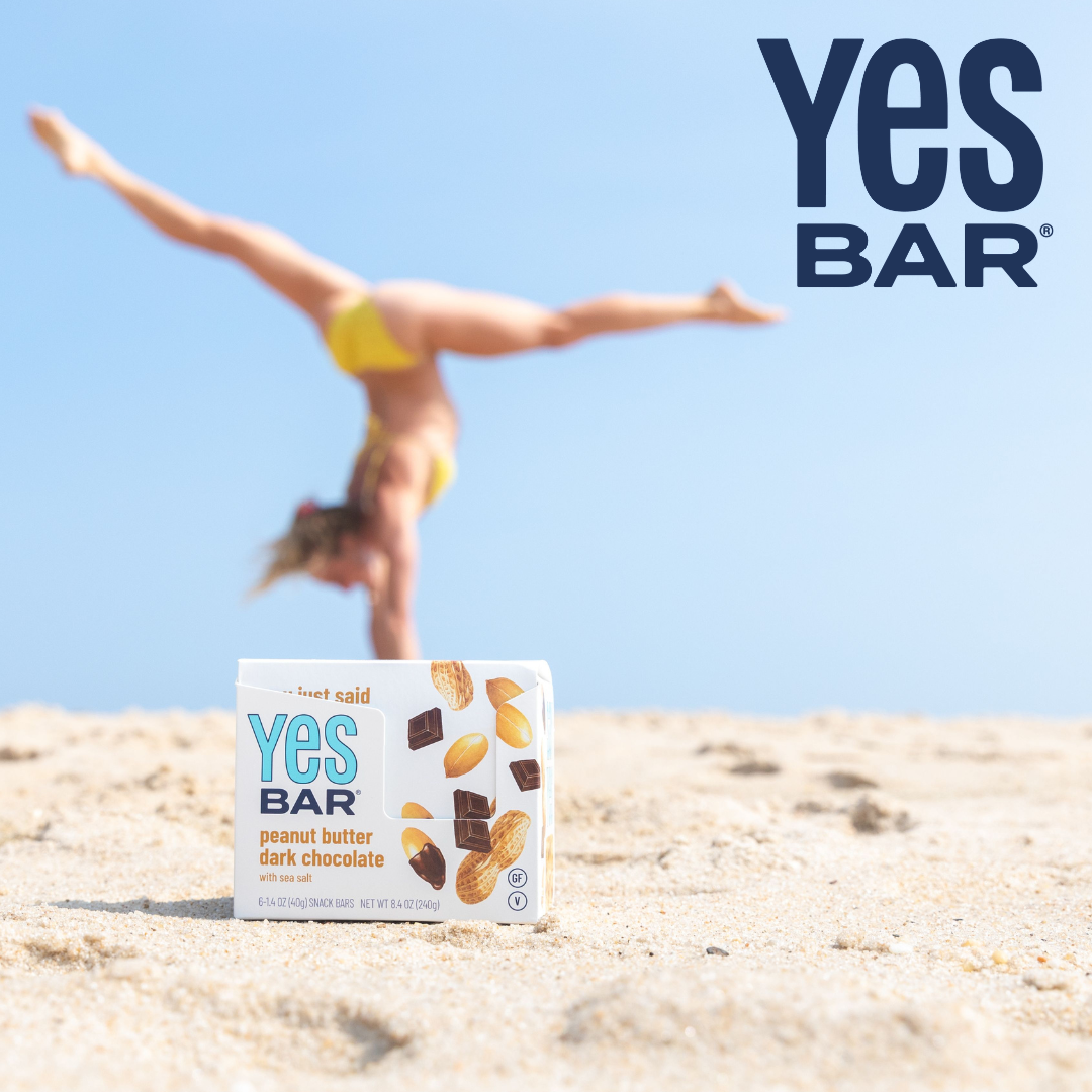 A Yes Bar snack box sits on sandy beach ground with a blurred woman balancing on her hands in the background under a blue sky. The Yes BAR logo is also shown in the upper right.
