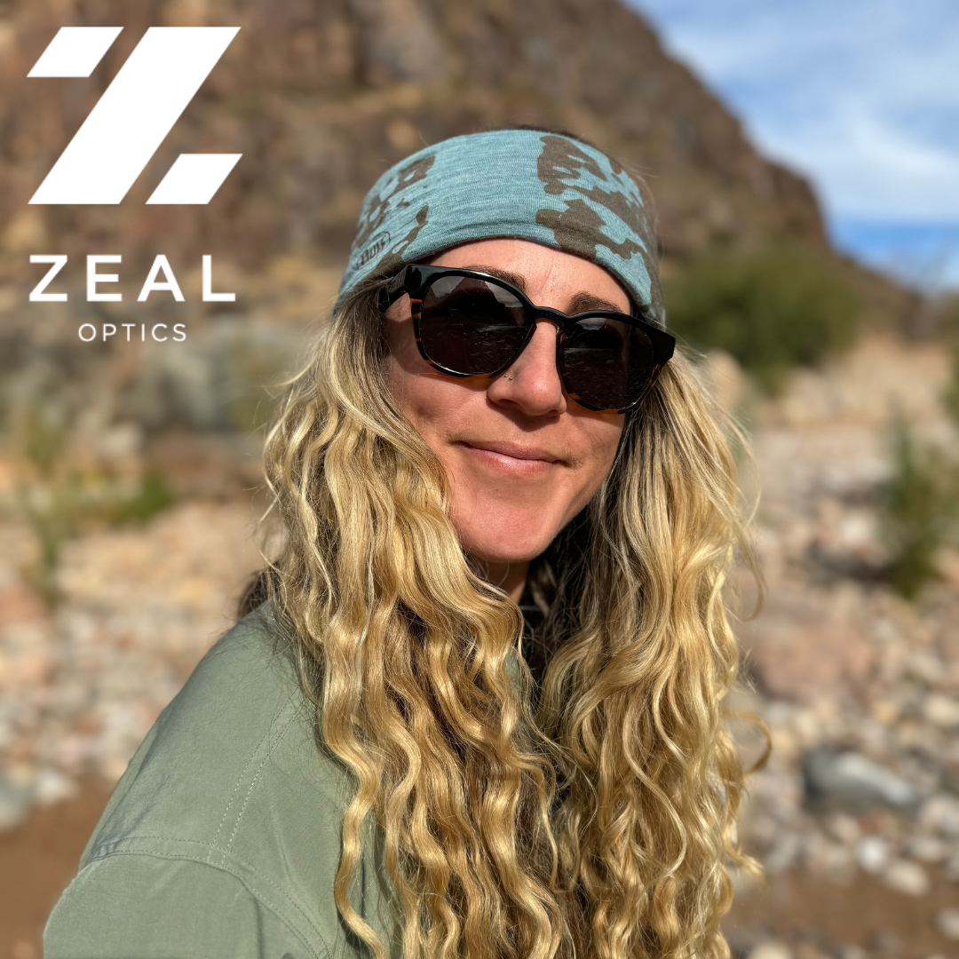 A woman outdoors wearing a teal camouflage Zeal Optics beanie, black sunglasses, and an olive green shirt, with a rocky mountain background and the Zeal Optics logo in the top left corner.