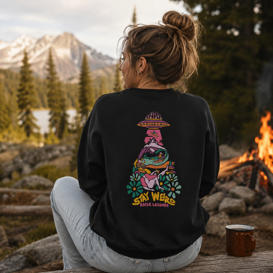 Person wearing a black sweatshirt with a colorful graphic and text, sitting by a campfire with mountains in the background.